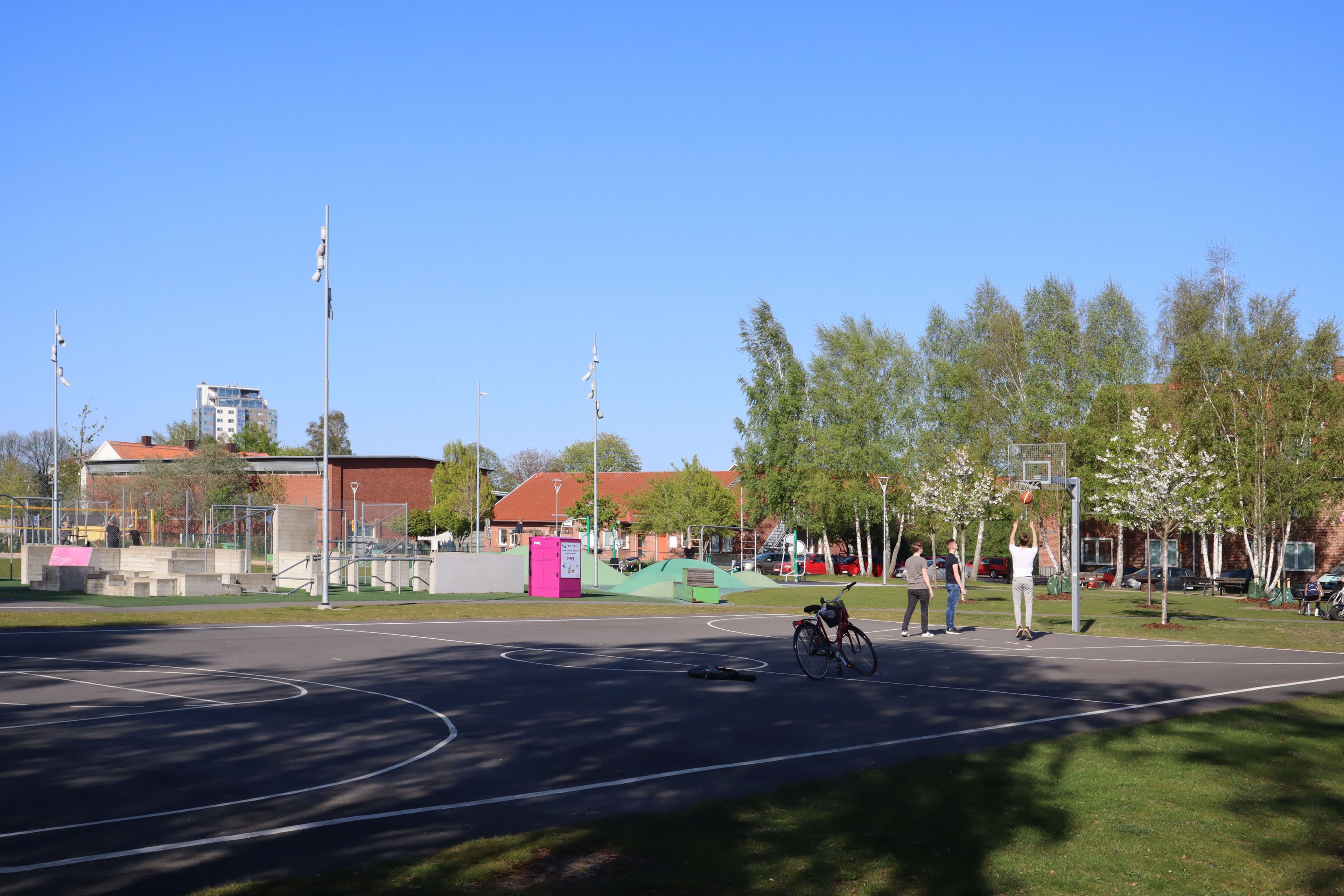 Tre killar spelar basket vid en Pifflbox i en park under en solig dag